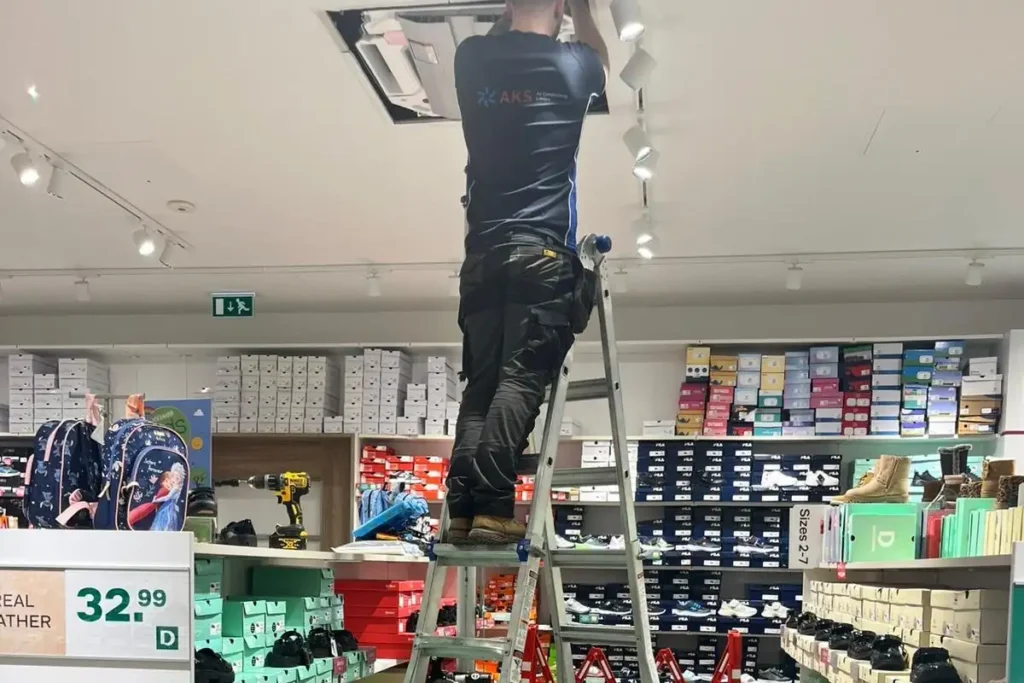 AKS Air Conditioning engineer working at height on a ceiling-mounted air conditioning unit inside a retail store, with safety barriers in place